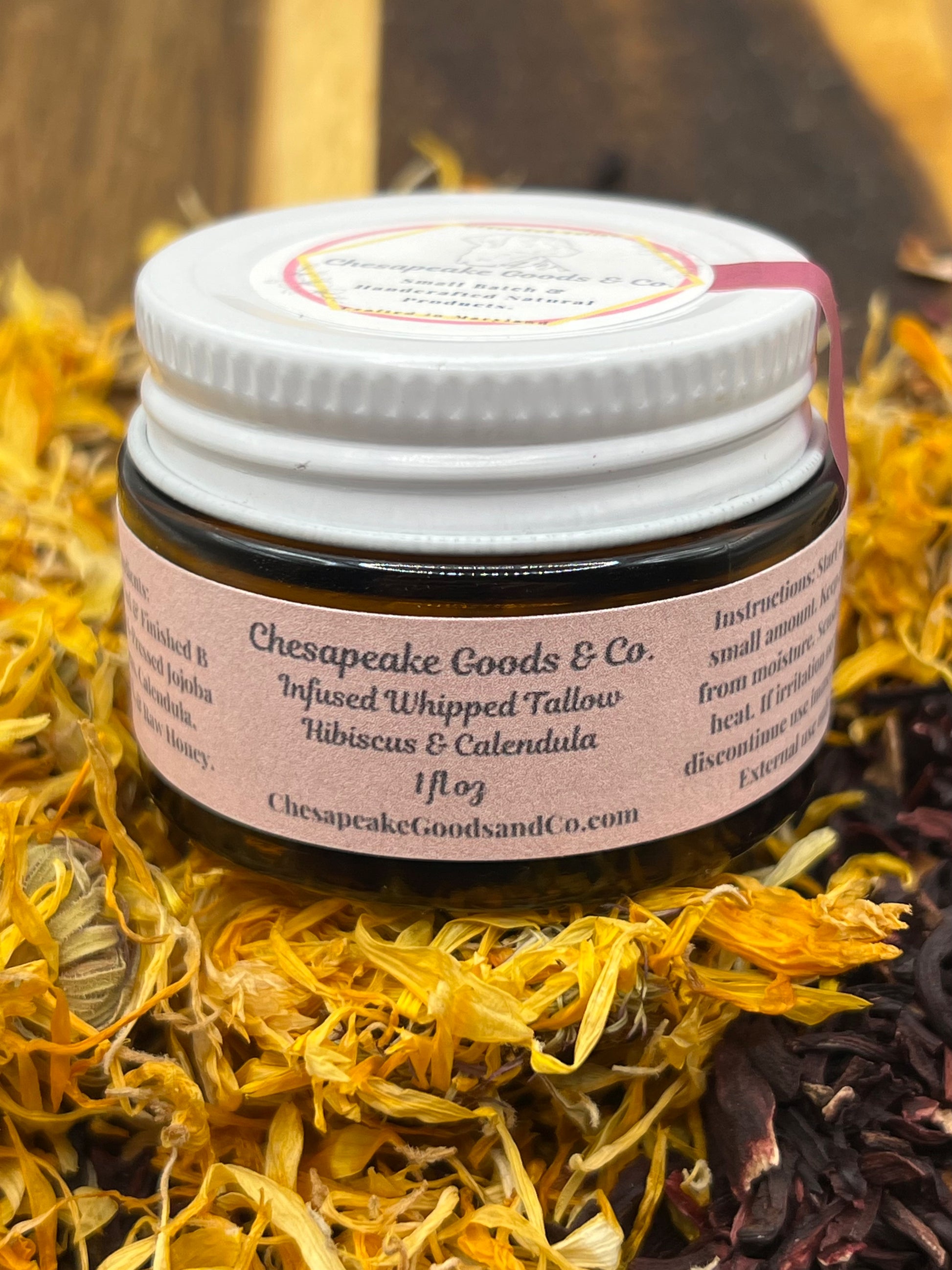 A 1 oz amber glass jar with pale pink label that says "Chesapeake Goods and Co, Infused Whipped Tallow Hibiscus and Calendula" with a white lid and tamper seal, surrounded by dried calendula and hibiscus petals on a wooden surface.
