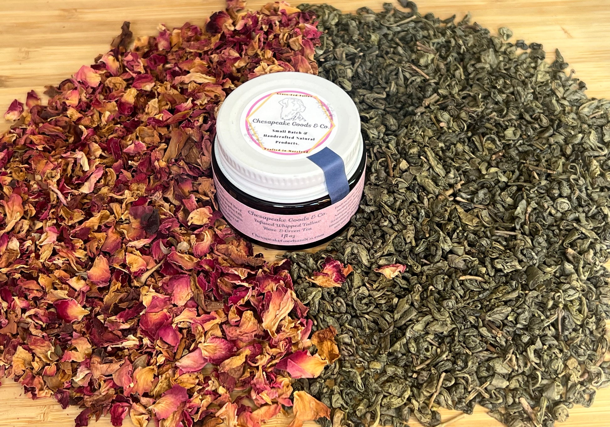 1 oz amber glass jar with a pink wrap label and white top, surrounded by dried green tea leaves and dried rose petals on a wooden surface.