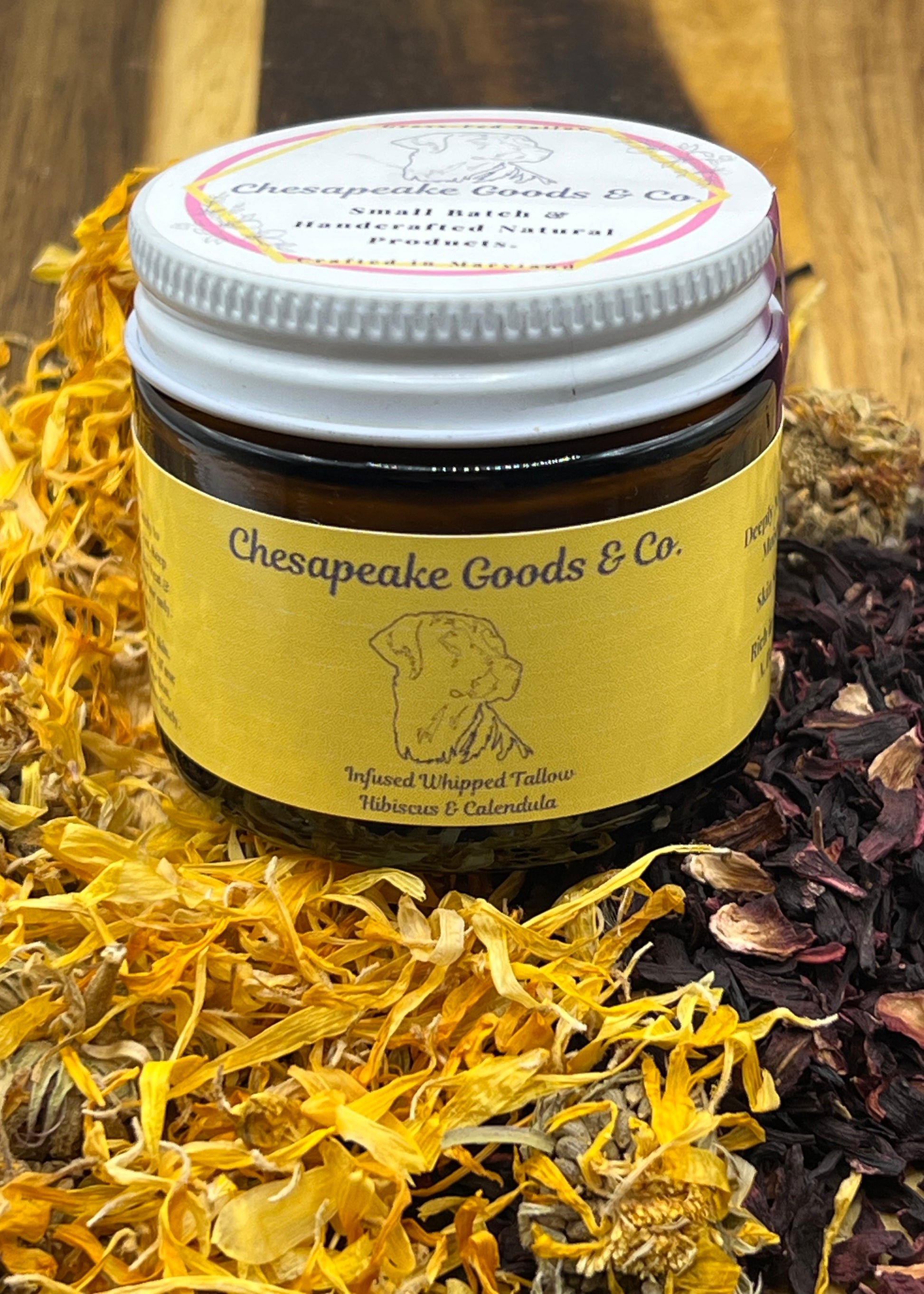 A 2 oz amber glass jar with a bright yellow wrap label that says "Chesapeake Goods and Co, Infused Whipped Tallow Hibiscus and Calendula" with a white lid surrounded by dried calendula and hibiscus petals on a wooden surface.

