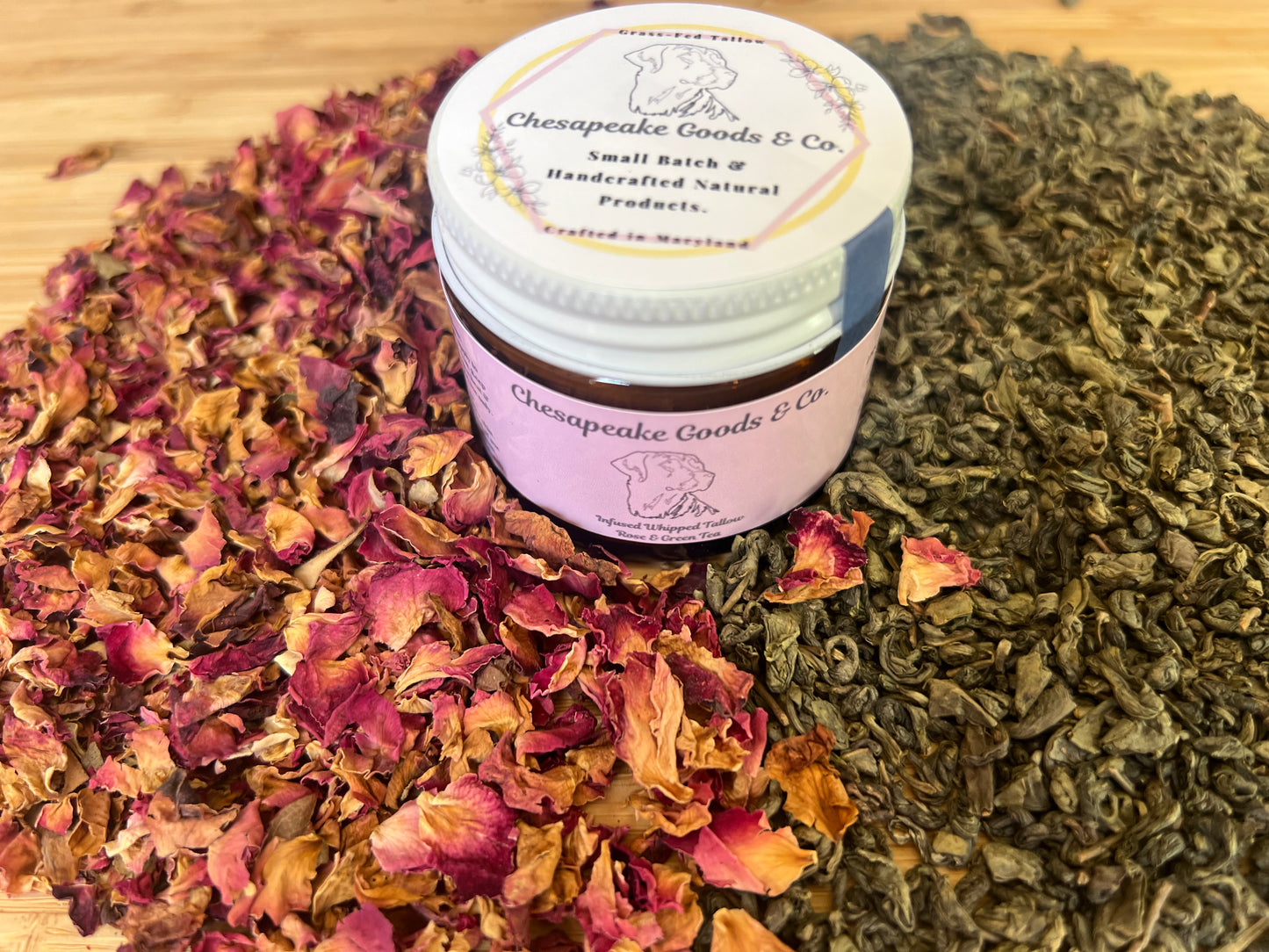 2 oz amber glass jar with pink wrap label that says "Chesapeake Goods and Co" with a white lid, surrounded by dried rose petals and green tea leaves.