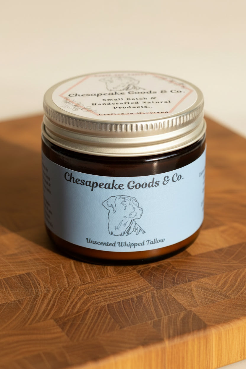 Chesapeake Goods & Co. sells unscented whipped tallow, a handcrafted natural product made in Maryland, packaged in a 2 oz amber glass jar with pale blue label sitting upon a wooden surface.
