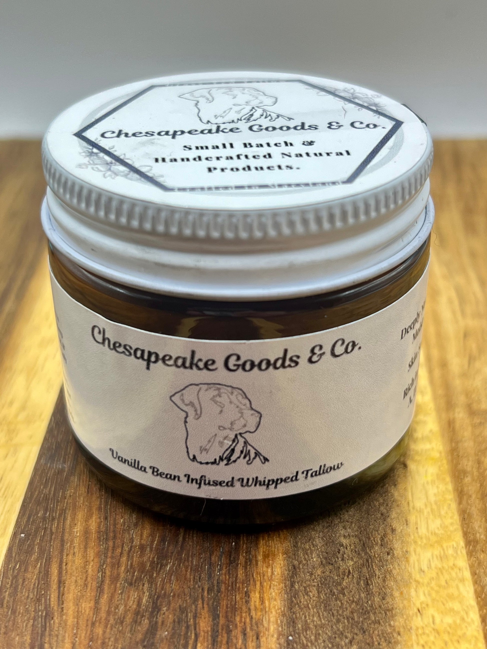 Amber 2 oz jar displays Chesapeake Goods & Co. Vanilla Bean Infused Whipped Tallow moisturizer on a wooden surface.
