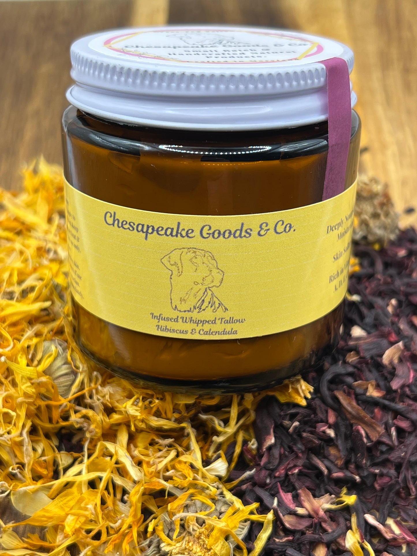 A 4 oz amber glass jar with a bright yellow lable that says "Chesapeake Goods and Co Infused Whipped Tallow, Hibiscus & Calendula" with a white lid and a pink tamper seal, surrounded by dried calendula and hibiscus petals on a wooden cutting board.  