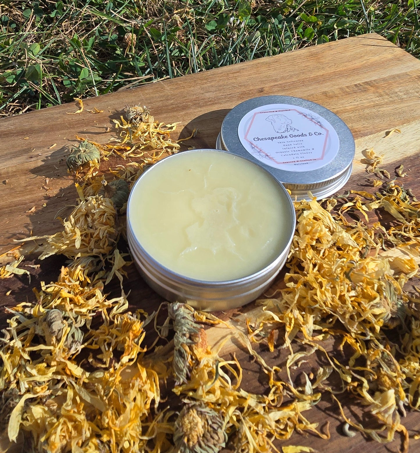Chesapeake Goods & Co. showcases their organic chamomile and calendula flower skin salve in a silver tin on a wooden board amongst scattered calendula flower petals.
