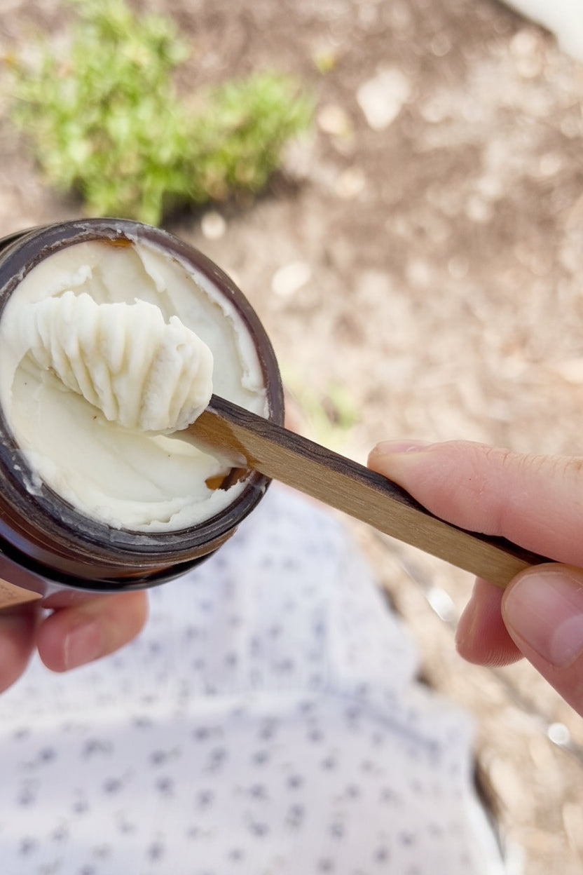 Person holding an amber glass jaw with a creamy balm inside, using an artisan wooden spatula to mix to show texture. 