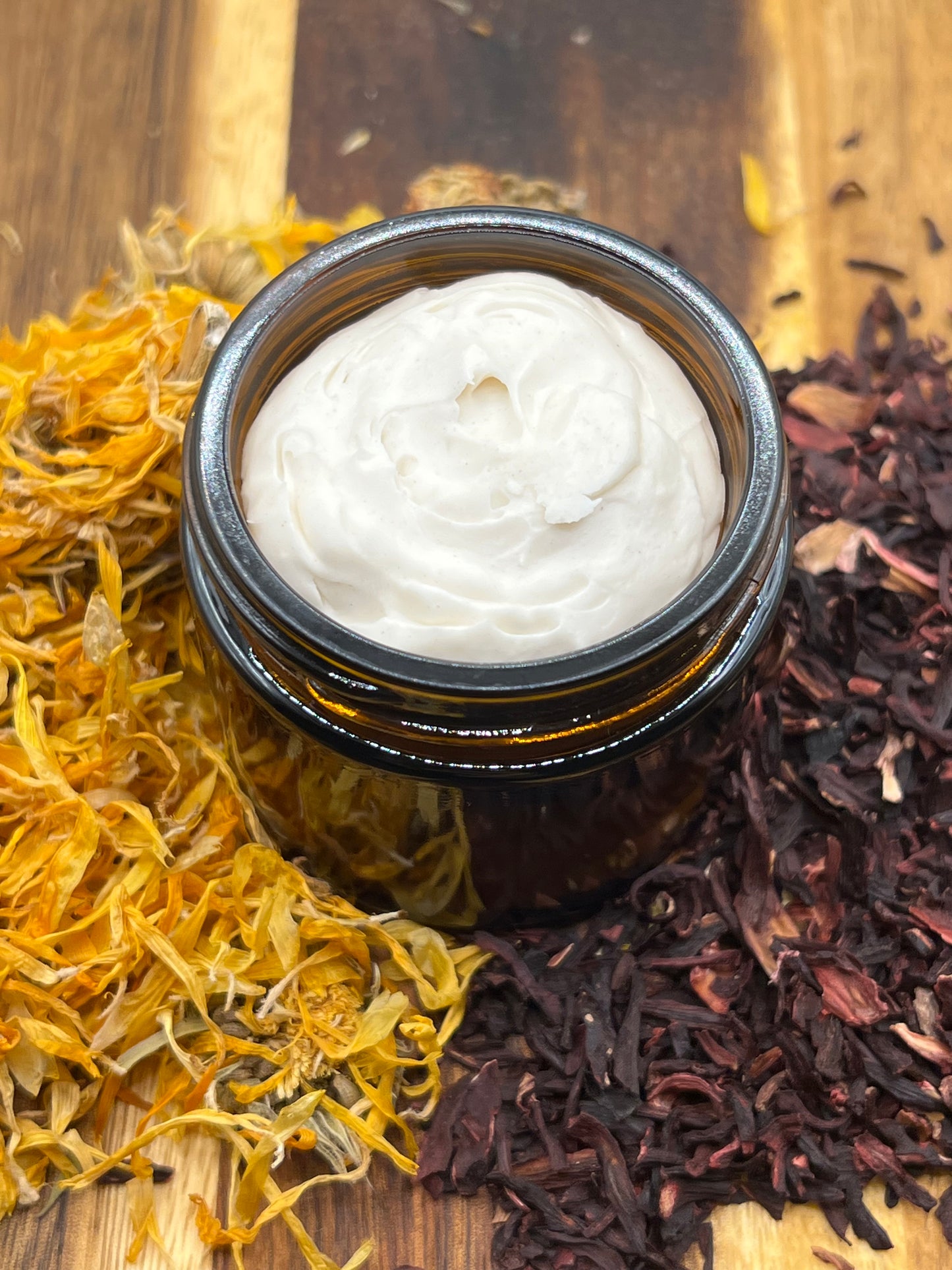 An amber jar of creamy tallow balm sits on a wooden surface surrounded by dried calendula petals and hibiscus.
