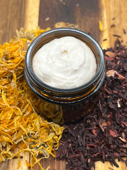 An amber jar of creamy tallow balm sits on a wooden surface surrounded by dried calendula petals and hibiscus.
