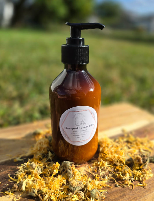 Chesapeake Goods & Co amber glass lotion pump bottle with a white circle label on the front sititng on a wooden surface with calendula flowers, outside in the grass.
