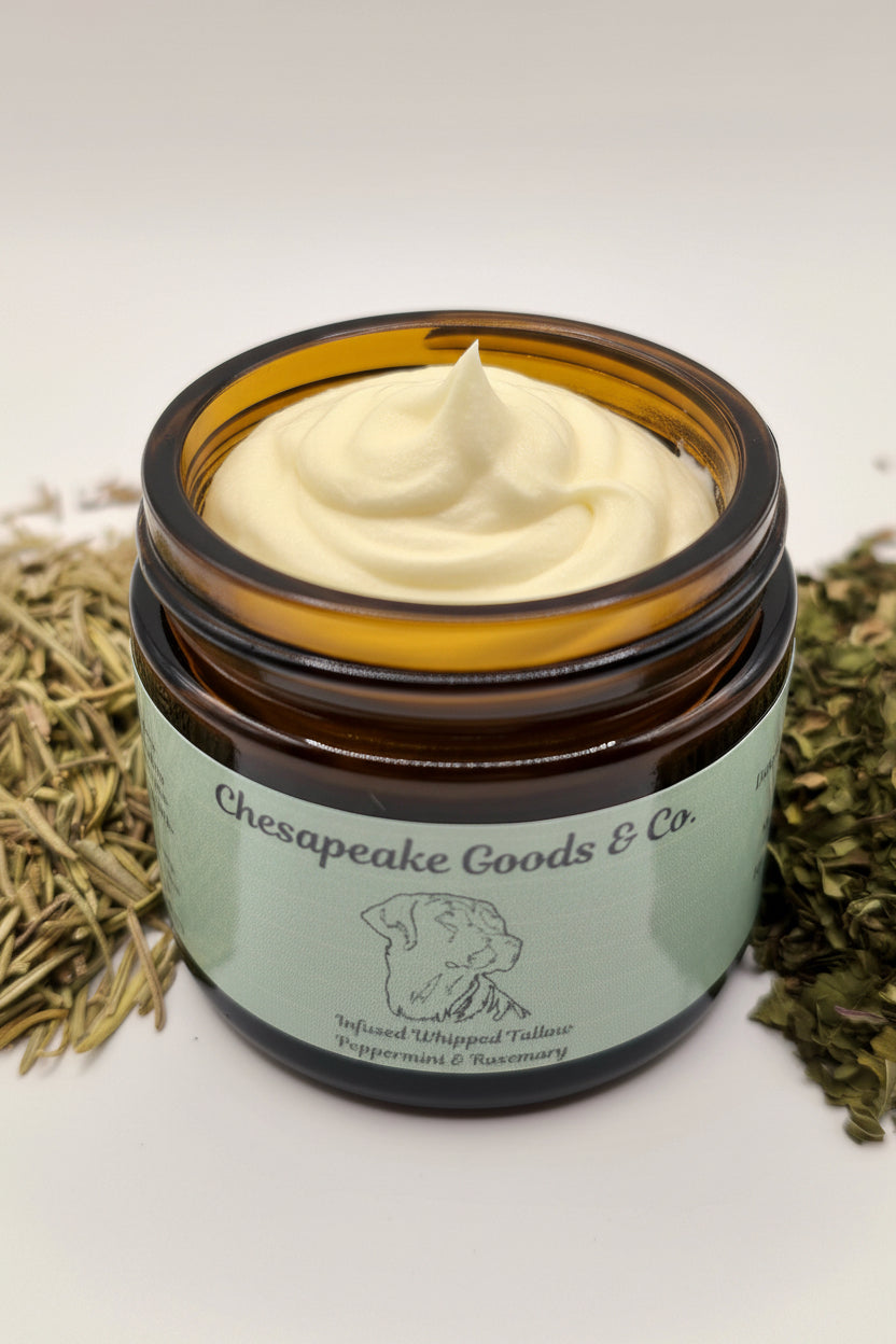 Chesapeake Goods & Co. whipped tallow balm, infused with peppermint and rosemary, sits open in a jar amidst dried peppermint and rosemary herbs.
