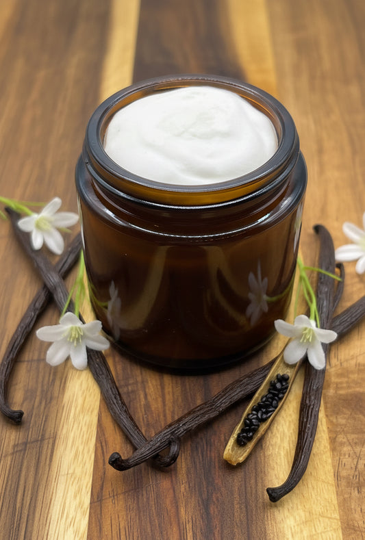 An amber glass jar of thick, white balm sits on a wood surface surrounded by vanilla bean and vanilla flowers.